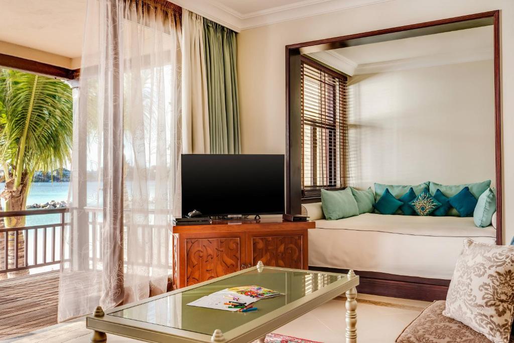 Mūsdienīgs viesnīcas numurs ar skatu uz jūru, kurā ir televizors, ērta sēdvieta ar spilveniem un lieli logi ar aizkariem.