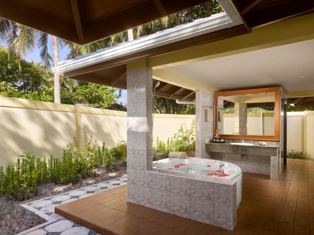 Mūsdienīga vannas istaba ar atvērtu dizainu, kurā ir vannu ar ziedlapiņām un plašu spoguli, ieskauta zaļiem augiem un saulainu ainavu.