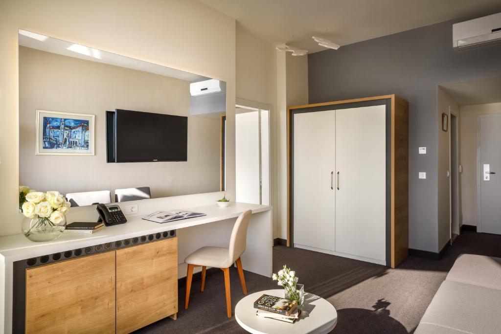 Mūsdienīga viesnīcas numura iekārtojums ar televizoru, darba vietu un skapi. Telpa ir gaiša un eleganta, ar neitrālām krāsām un dekoratīvām detaļām.