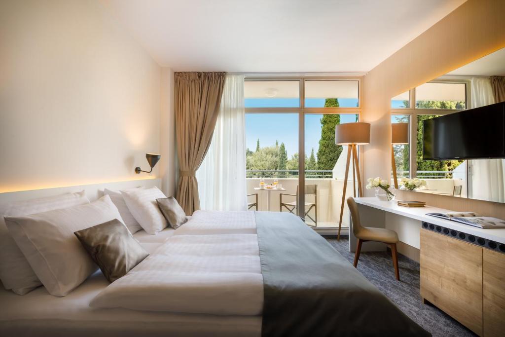 Mūsdienīga viesnīcas numura iekārtojums ar divām gultām, televizoru un skatu uz balkonu, kurā redzams dārzs un zaļumi. Numurā ir gaismas un stilīgi dekorēti elementi, radot mājīgu atmosfēru.