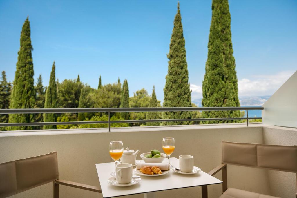 Balkons ar skatu uz zaļām priedēm, uz galda novietots brokastu komplekts - glāze sulas, kafija, augļi un kruasāni.