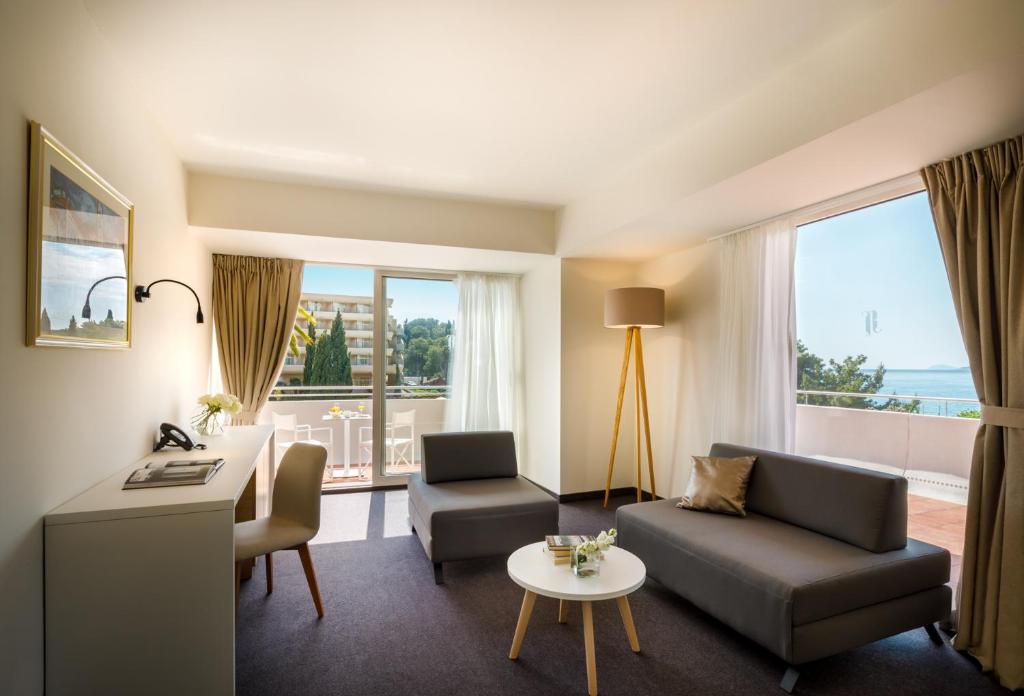 Mūsdienīgs viesnīcas numurs ar skatu uz jūru, gaišām sienām un minimālistisku interjeru, kurā ir dīvāns, galdiņš un darba stacija.