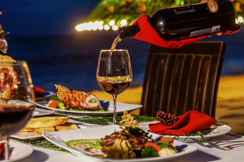 Romantisks vakariņu galds pie pludmales, kur pasniedz sarkanvīnu un izsmalcinātus ēdienus, radot patīkamu atmosfēru. Gaismas fons un dabiskais skaistums piešķir burvību šai vakariņām.