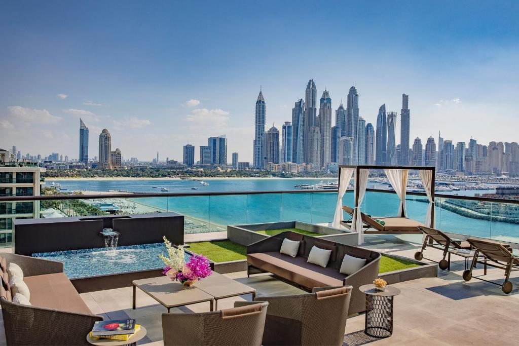 Mūsdienīgas terases skats uz Dubaijas pilsētas ainavu, kur redzami augstceltnes un jūra. Terasē ir mēbeles atpūtai un dekoratīvi augi, kas rada relaksējošu atmosfēru.