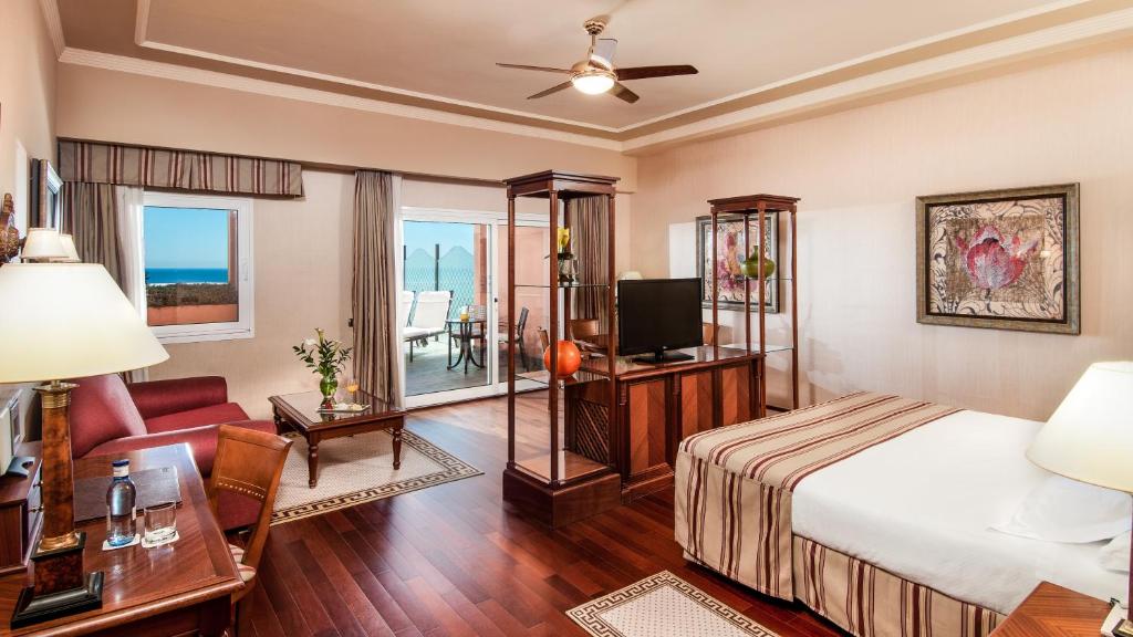 Elegants viesnīcas numurs ar skatu uz jūru, koka grīdu un mūsdienīgu iekārtojumu, kas ietver televizoru un ērtu atpūtas zonu. Gaisma un telpas plašums rada patīkamu atmosfēru.