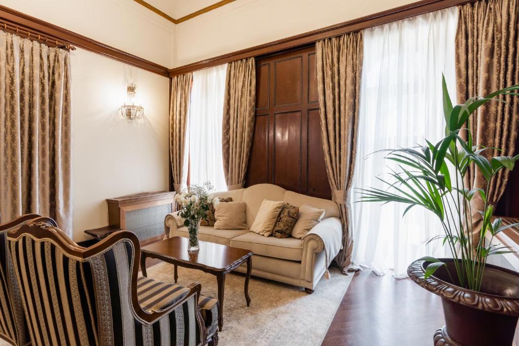 Eleganta viesistaba ar mīksto dīvānu, vintage stila krēsliem un greznu aizkaru. Telpu papildina eleganta auga pods un gaismas avots, radot siltu un viesmīlīgu atmosfēru.