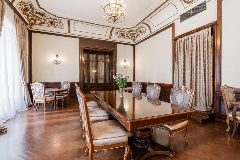 Eleganta ēdamzāle ar greznu koka galdu un izsmalcinātiem krēsliem, kuriem ir mīkstas sēdekļi. Telpu papildina grezns lustras apgaismojums un dekoratīvi logi, radot patīkamu atmosfēru.