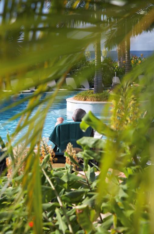 Relaksējoša ainava pie baseina ar palmām un zaļumiem, kurā redzams cilvēks, kas atpūšas uz krēsla. Ideāla vide atpūtai un saulē.