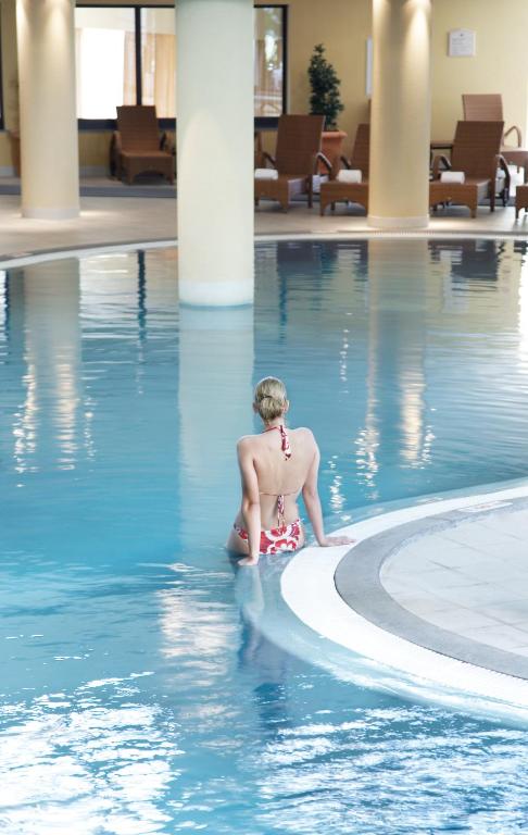 Sieviete sarkanā peldkostīmā sēž pie baseina, baudot mierīgu atmosfēru luksusa spa vidē. Apkārt ir gaišas krāsas interjers un ērtas atpūtas zonas.
