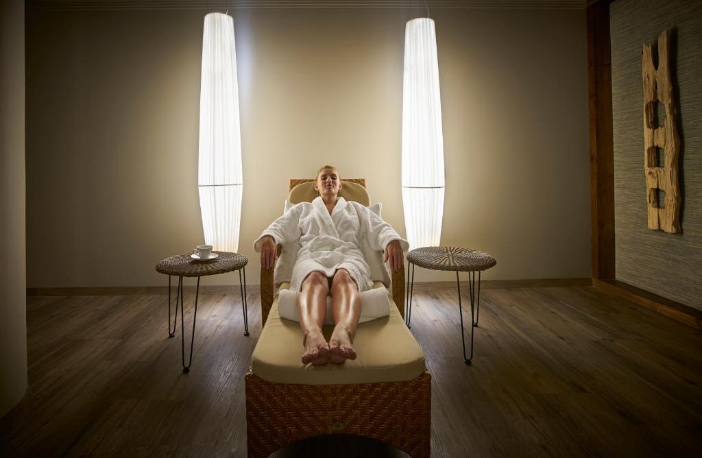 Relaksējoša spa atmosfēra ar personu, kas ģērbta baltā halātā, sēžot uz ērta krēsla. Apkārt ir eleganta apgaismojuma un koka akcenti.