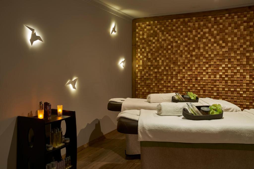 Mūsdienīgs spa interjers ar koka sienu un relaksējošu atmosfēru, kurā redzami masāžas galdi un sveces. Gaismas efekti piešķir telpai mierīgu noskaņu.