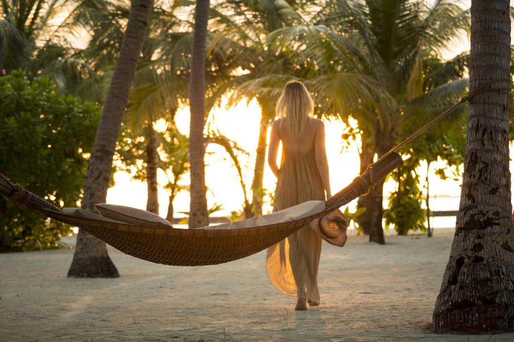 Sieviete, tērpta vieglā kleitā, pastaigājas gar šūpuļtīklu pludmalē, apņemta palmām un saulrieta gaismas.