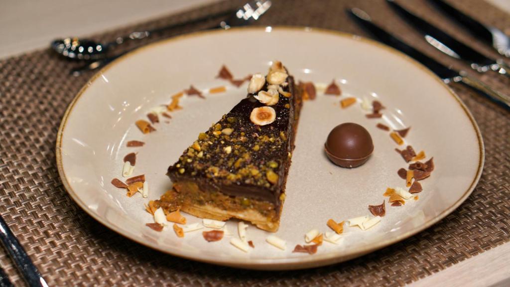 Dekoratīvs deserts ar riekstiem un šokolādi, pasniegts uz elegantas šķīvja, rotāts ar sagrieztiem mandelēm un šokolādes gabaliņiem.