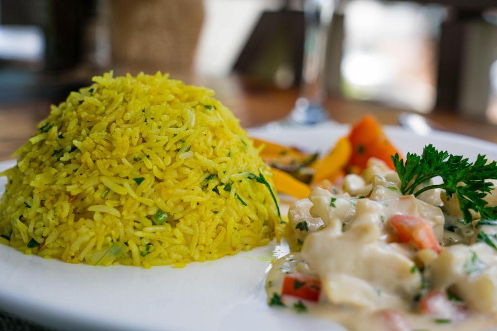 Dzeltenās rīsu porcijas ar dārzeņiem un krēmīgu mērci, pasniegtas uz baltā šķīvja.