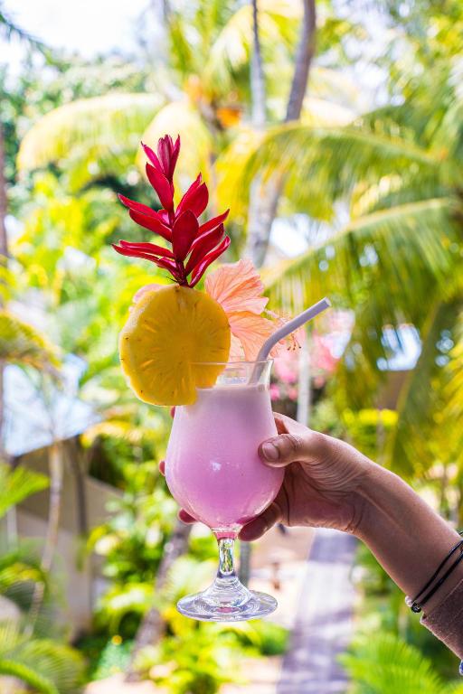 Svaigs un krāsains kokteilis ar ananāsu un ziedu rotājumu, fona attēlā redzamas palmas un tropu vide.