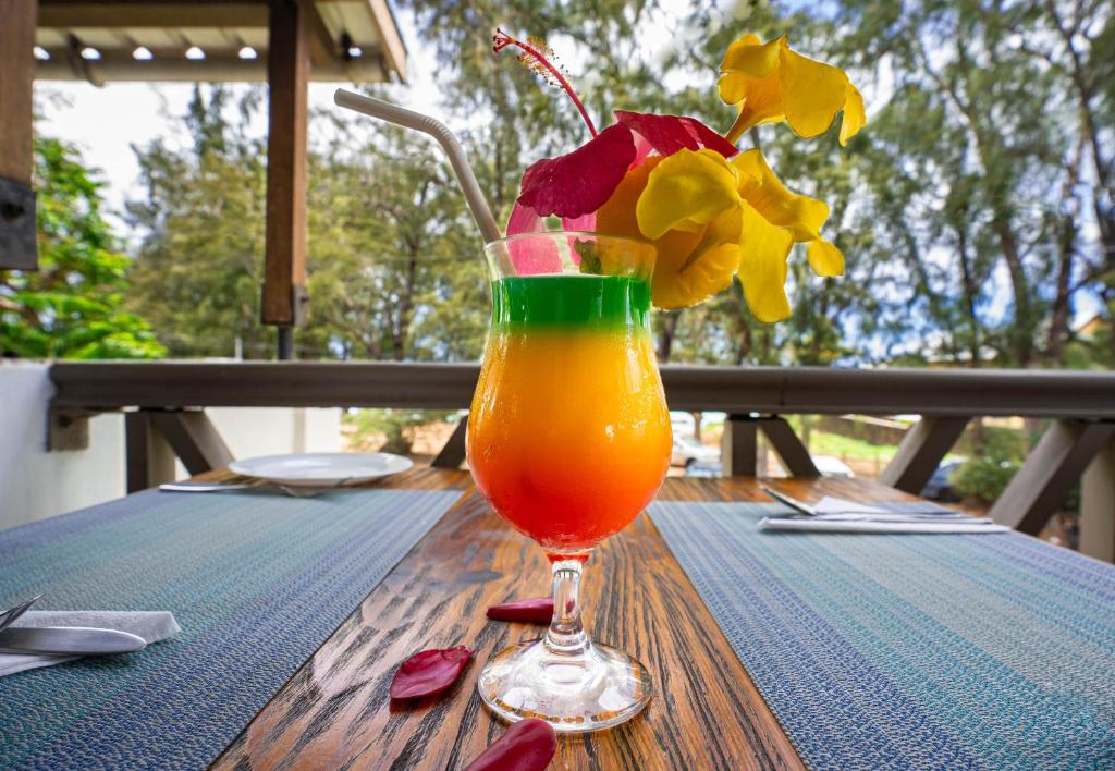 Krāsains kokteilis ar zaļu, dzeltenu un sarkanu slāni, rotāts ar ziediem, novietots uz koka galda. Fons attēlo dabu un ērtu ēdamstāvu.