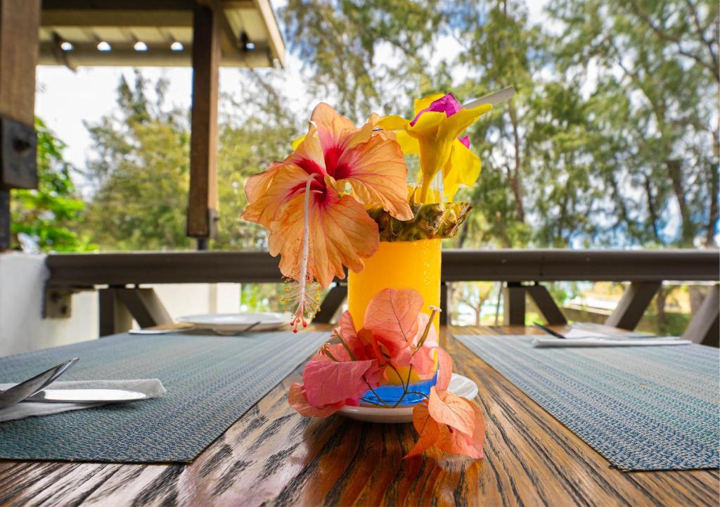 Kokteilis ar krāsainām ziedu dekorācijām uz koka galda, fons ar zaļiem kokiem un gaišu debesi.