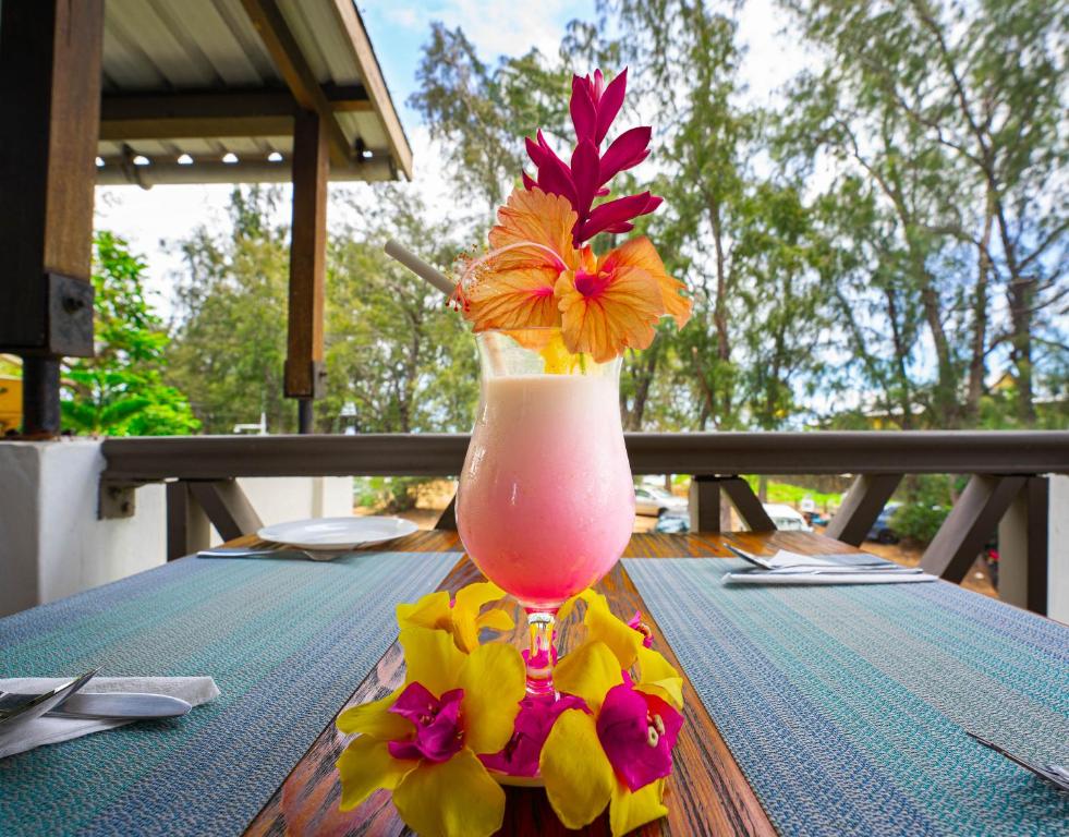 Eksotiska kokteiļa glāze ar ziediem uz galda, kas atrodas ārā pie restorāna. Fonā redzamas zaļas koku lapas un skaidrs debesis.