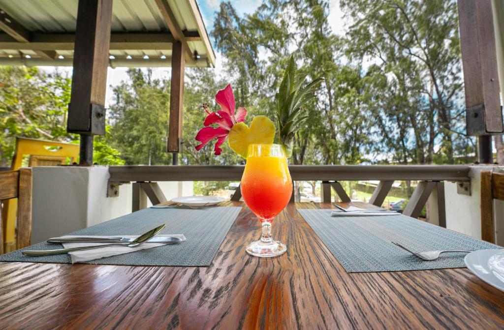 Tropisks kokteilis ar rotājumiem no ziediem un ananāsa, pasniegts uz koka galda restorāna terasē, ar zaļojošiem kokiem fonā.