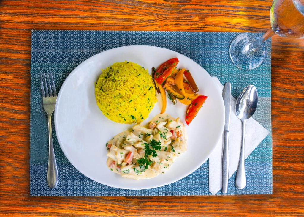 Gards ēdiens uz balta šķīvja, kurā ir dzeltenā rīsa porcija, krēmīga mērce ar dārzeņiem un cepti tomāti. Fonā ir galds ar galdautu un glāzi ūdens.