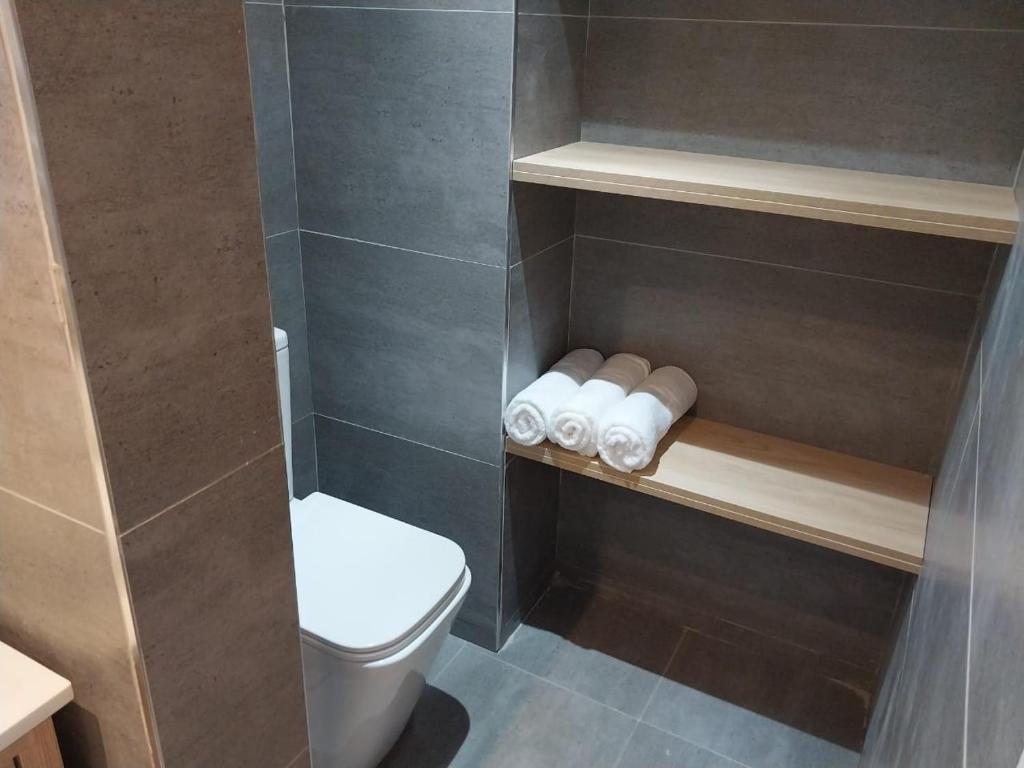 Mūsdienīgs vannas istabas interjers ar pelēkām flīzēm, tualeti un koka plauktu, uz kura novietoti salocīti dvieļi.