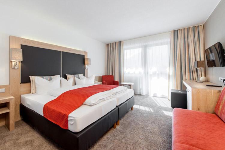 Mūsdienīga viesnīcas istaba ar dubultgultu, sarkanu segu un ģimenes telpu. Telpa ir gaiša ar lieliem logiem un modernu dizainu.