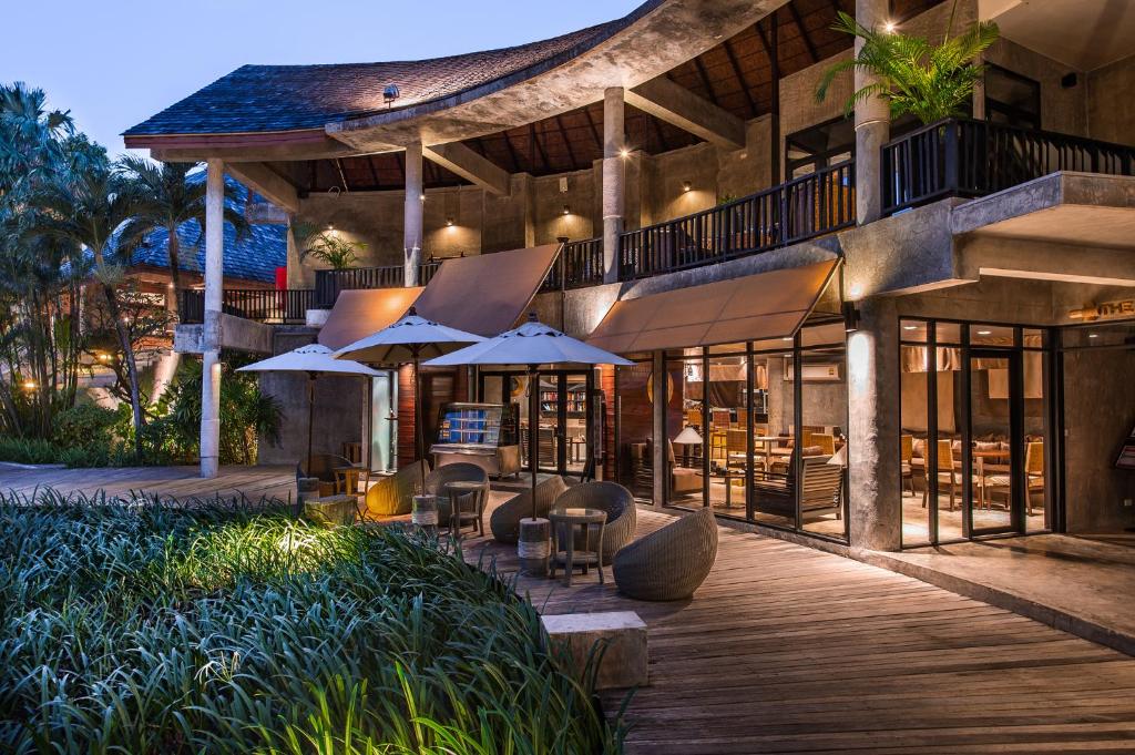 Mūsdienīgs viesnīcas ārpuses skats ar terasi, kurā ir sēžamie krēsli un saulessargi, apzaļumots dārzs un ēka ar interjera apgaismojumu.