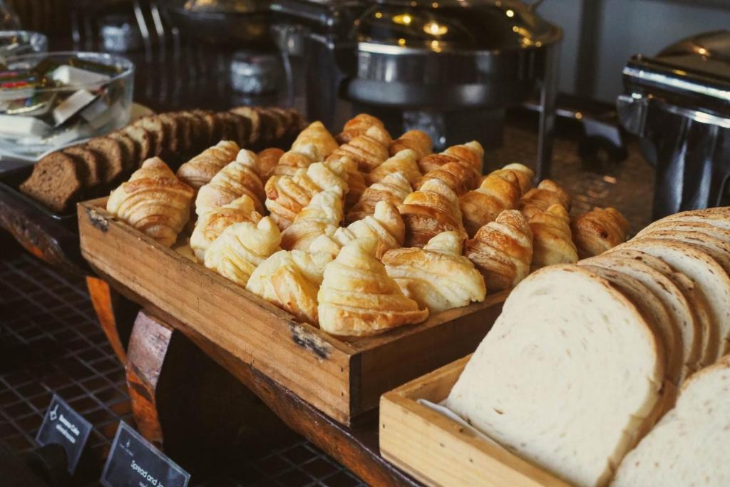 Svaigi cepti kruasāni un maizes šķēles izkārtoti koka traukā, blakus citiem konditorejas izstrādājumiem.