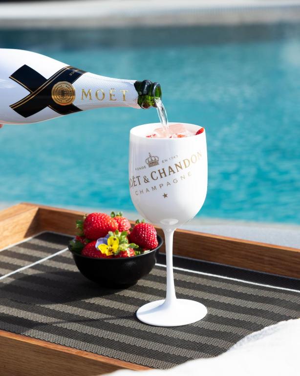 Šampanieša pudele tiek ielejta elegantā glāzē ar ledus un zemenēm, blakus atrodas augļu bļoda, viss novietots uz galda pie baseina.