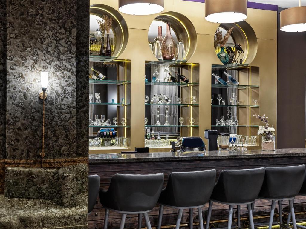 Mūsdienīgs bārs ar elegantiem apgaismojuma elementiem un stilīgām glāzēm, izvietots pie pievilcīgas zelta plaukta. Interjers apvieno dabīgus materiālus un modernu dizainu.