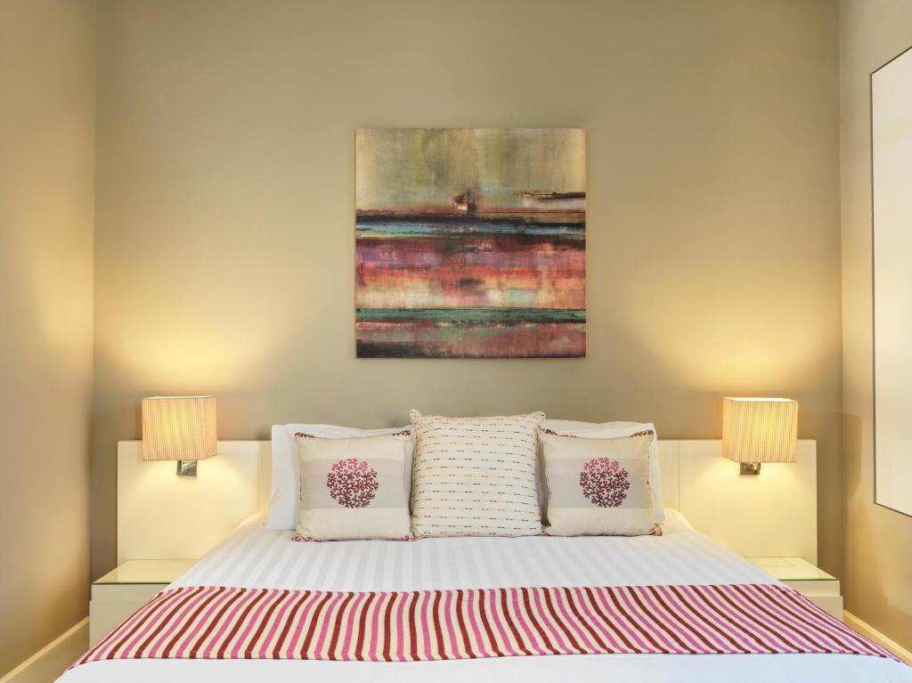 Mūsdienīgs guļamistabas interjers ar gultu, dekoratīvām spilvenēm un mīkstām lampām abās pusēs. Uz sienas ir krāsaina gleznas fragmenta dekorācija, kas piešķir telpai siltumu un stilu.