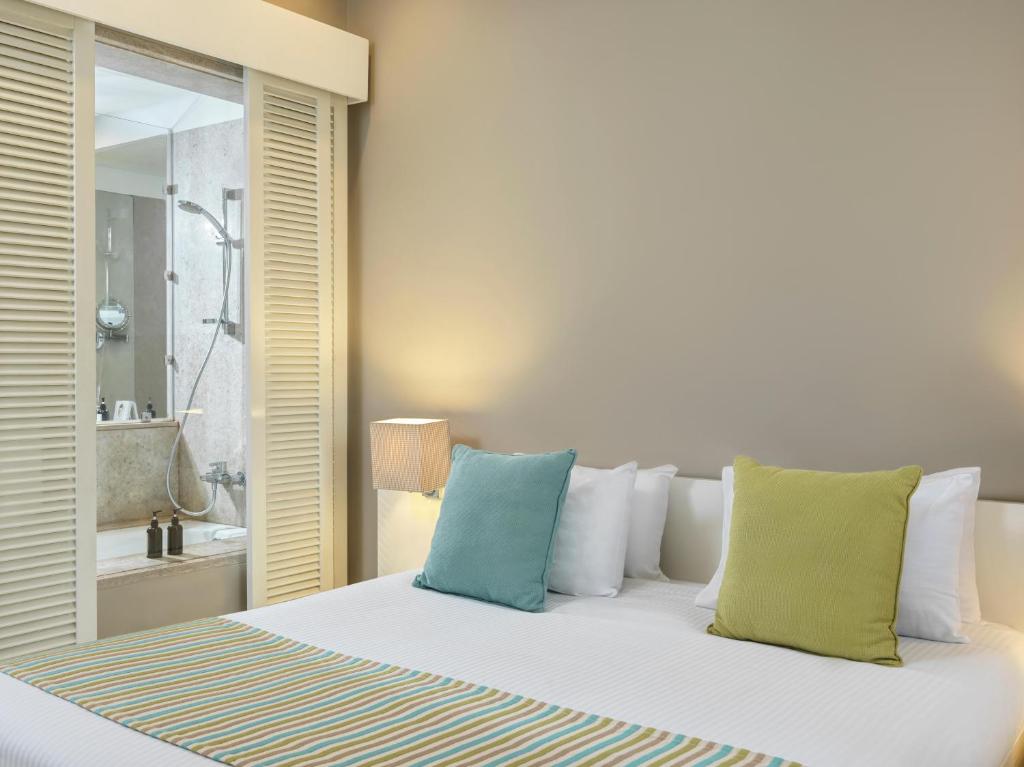 Mūsdienīgs guļamistabas interjers ar gultu, spilveniem un skatu uz vannas istabu caur stikla durvīm. Gaismas un krāsainas akcenti piešķir telpai siltumu un mājīgumu.