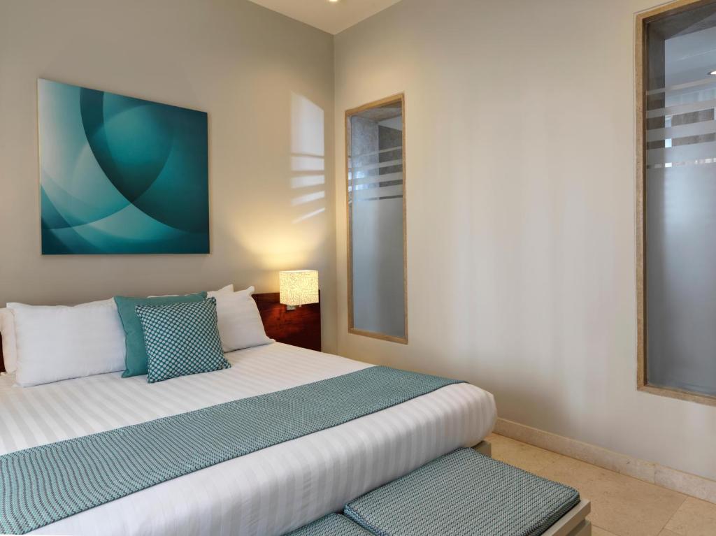 Mūsdienīgs guļamistabas interjers ar gultu, dekorētu ar zilu un balto tekstilu, un abstraktu mākslas darbu uz sienas. Telpa ir gaiša un eleganta, radot relaksējošu atmosfēru.