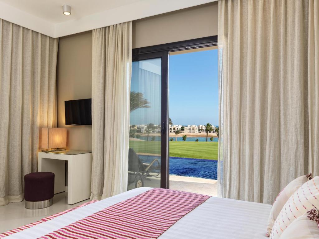 Mūsdienīga guļamistaba ar panorāmas skatu uz dārzu un peldbaseinu, iekārtota ar televizoru un elegantiem aizkariem. Gaisma plūst cauri logiem, radot patīkamu atmosfēru.