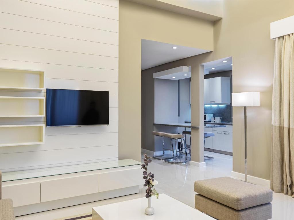 Mūsdienīgs viesistabas interjers ar televizoru uz sienas, atvērtu virtuvi un ērtu dīvānu. Gaismas tonis un minimalistisks dizains rada patīkamu atmosfēru.