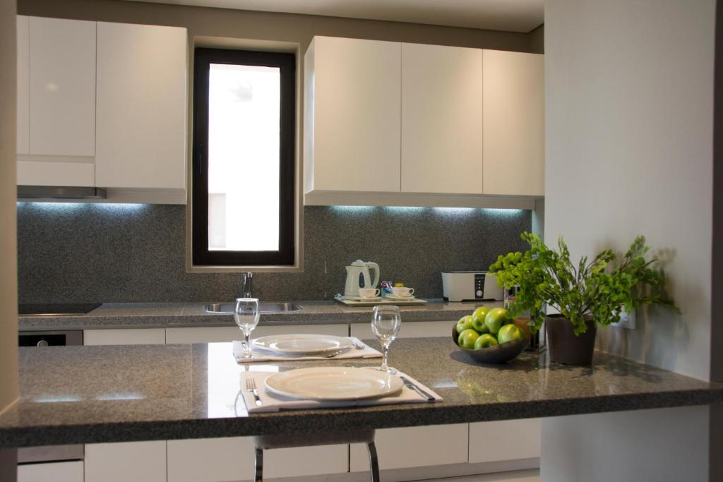 Mūsdienīga virtuve ar pelēku darba virsmu, baltām skapīšiem un logu, caur kuru ieplūst dabiska gaisma. Uz galda ir šķīvji, glāzes un augļu trauks ar zaļām āboliem.