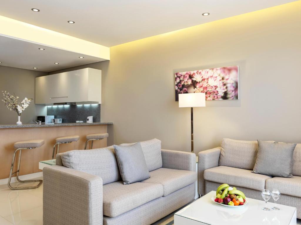 Mūsdienīga viesistaba ar pelēku dīvānu, baltiem mēbeļu akcentiem un modernu virtuvi. Uz galda ir augļu trauks un dekoratīva lampa, radot mājīgu atmosfēru.