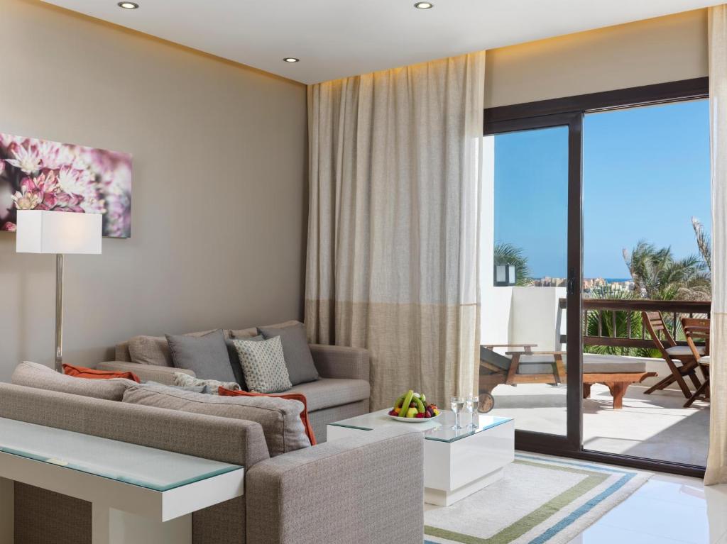 Mūsdienīga viesnīcas viesistaba ar gaišiem toņiem, mīkstām mēbelēm un lieliem logiem, kas piedāvā skatu uz āru. Galds ar augļiem un atpūtas zona uz balkona.