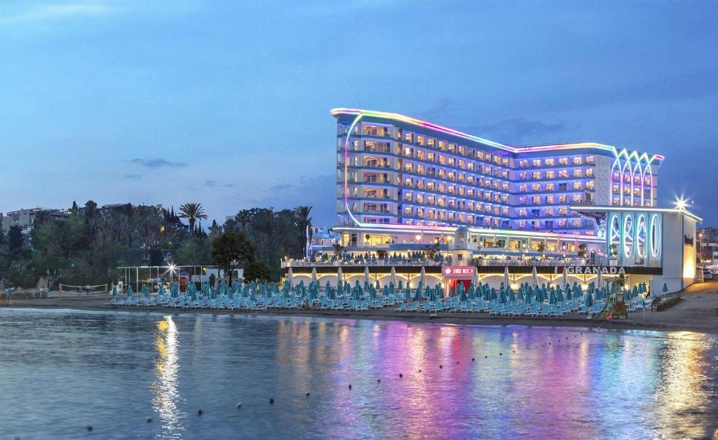 Mūsdienīgs viesnīcas komplekss pie pludmales ar krāsainu apgaismojumu, kas izgaismo ēku vakarā. Viesnīca atrodas pie ūdens ar pludmales sauļošanās krēsliem priekšā.