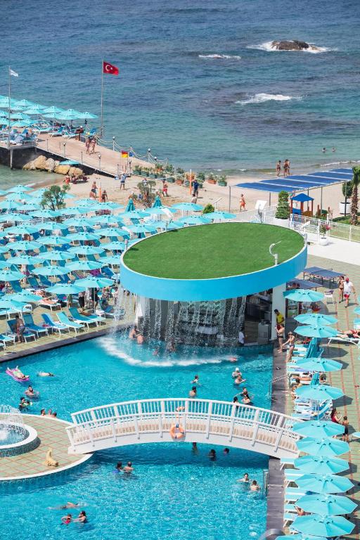 Vasaras kūrortā redzama peldbaseina zona ar zilu ūdeni, apņemta ar saulessargiem un sauļošanās krēsliem. Fons veido pludmale un jūra, bet redzama arī ūdens strūklaka un cilvēki, kas izbauda atpūtu.