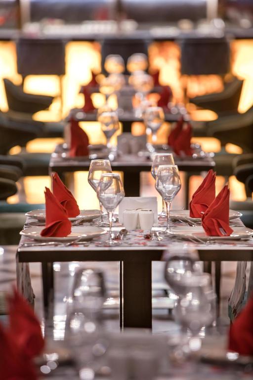 Eleganti iekārtota restorāna galdi, ar sarkaniem salvetēm un glāzēm, izveidojot viesmīlīgu atmosfēru. Galdi ir sakārtoti rindā, radot harmonisku noskaņu.