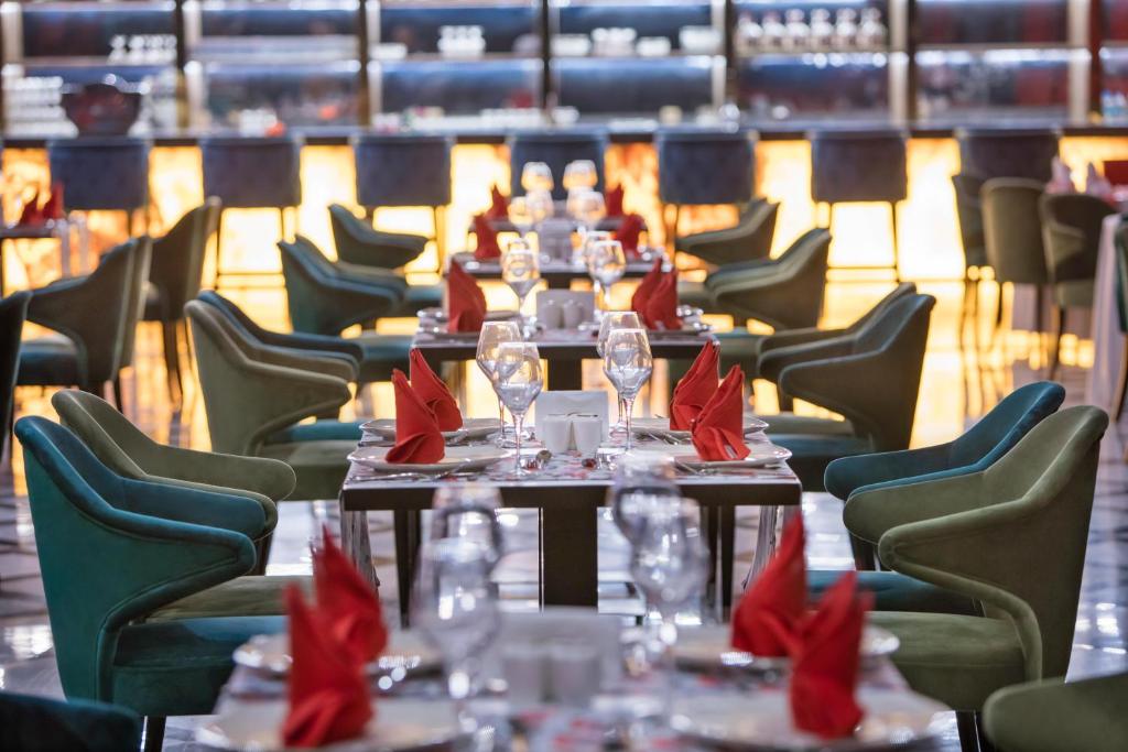 Elegants restorāna interjers ar zaļām un tumši zilu krēslu rindām, galdiem ar sarkanām servetēm un glāzēm. Gaisma rada siltu atmosfēru, aicinot uz patīkamu maltīti.