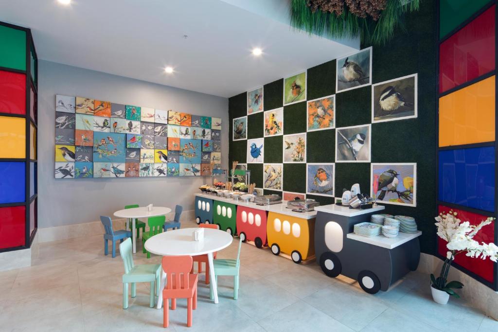 Krāsaina bērnu atpūtas telpa ar apkalpošanas letēm, kas izskatās kā vilcieni, un putnu mākslas darbiem uz sienām. Pie galdiem ir krāsainas bērnu krēsli.