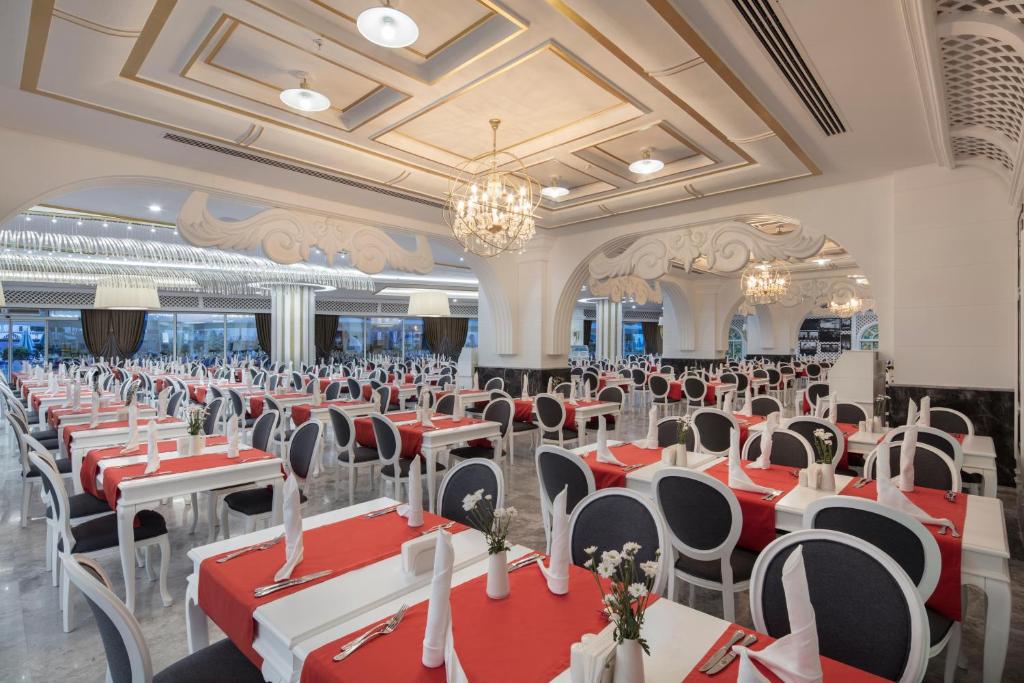 Elegants restorāns ar baltām galdiem un sarkaniem galdautiem, kas ir izrotāti ar ziediem. Telpa ir plaša, ar greznām detaļām un modernu apgaismojumu.