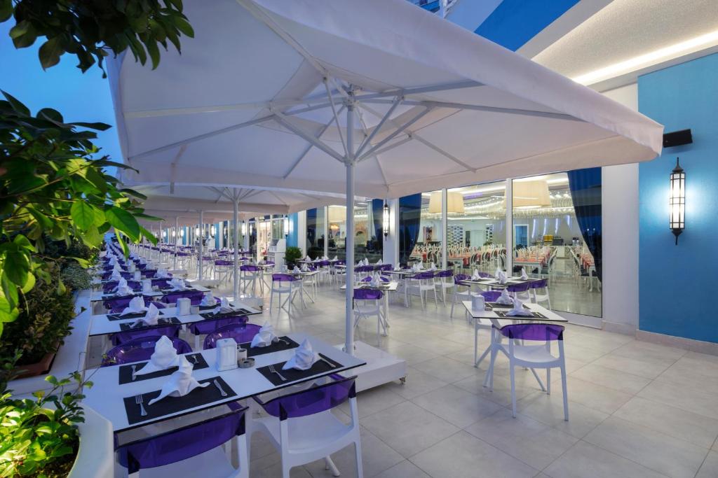 Āra restorāna terase ar baltām galdiem un violetām detaļām, kas izkārtoti zem baltiem saules aizsargiem, radot relaksējošu atmosfēru. Fons ietver modernu iekštelpu, kas ir redzama caur stikla durvīm.