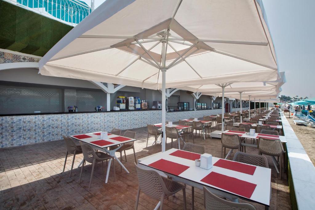 Saulaina pludmales kafejnīca ar terasi, kurā ir elegantas galds un krēsli, pārklāti ar baltiem saulessargiem. Fons parāda pludmales atmosfēru ar smiltīm un sauļošanās krēsliem.