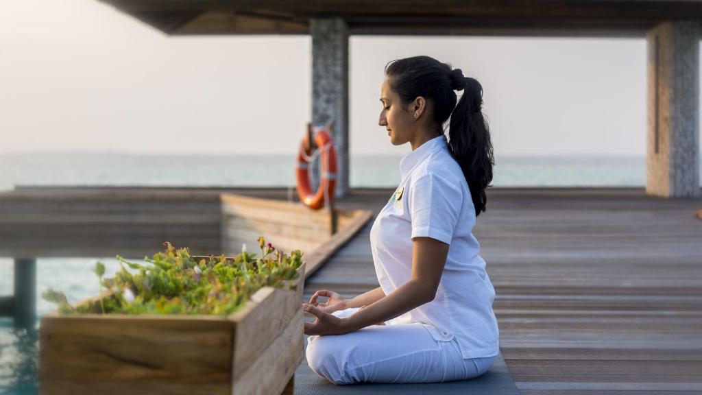 Sieviete mierīgā meditācijas pozīcijā pie ūdens, ar zaļām augu kompozīcijām priekšā. Rāmā vide veicina relaksāciju un garīgu atjaunošanos.