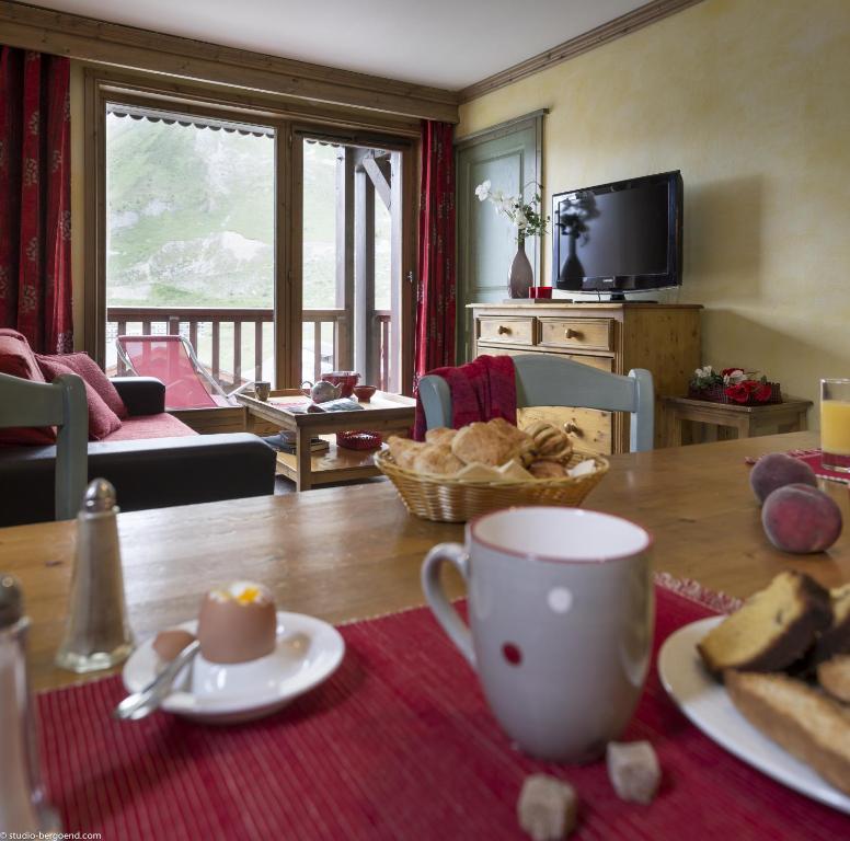 Mājas interjers ar koka mēbelēm, ēdamgaldu un brokastu galdu, kas klāts ar maizi un dzērieniem, skats uz kalniem caur logiem.