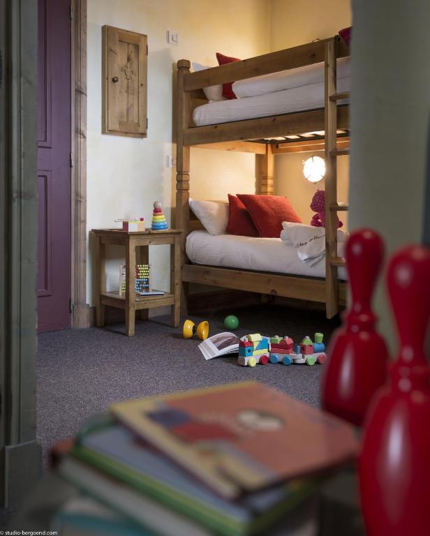 Bērnu istaba ar divstāvu gultu, kurā ir spilveni un rotaļlietas. Uz grīdas redzami krāsaini kluču komplekti un spēļu priekšmeti, kas liecina par aktīvu bērnu klātbūtni.
