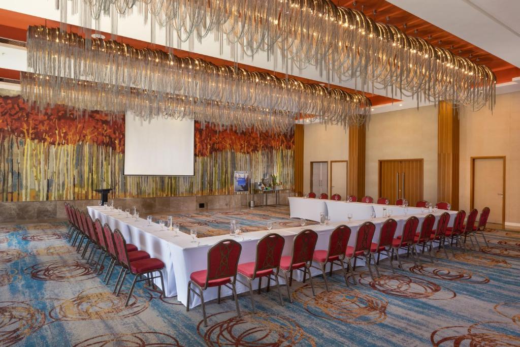 Konferenču zāle ar elegantām sarkanām krēslām un izsmalcinātu lampu, dekorēta ar krāsainu sienu gleznojumu. Iekārtojums paredzēts sanāksmēm un pasākumiem.